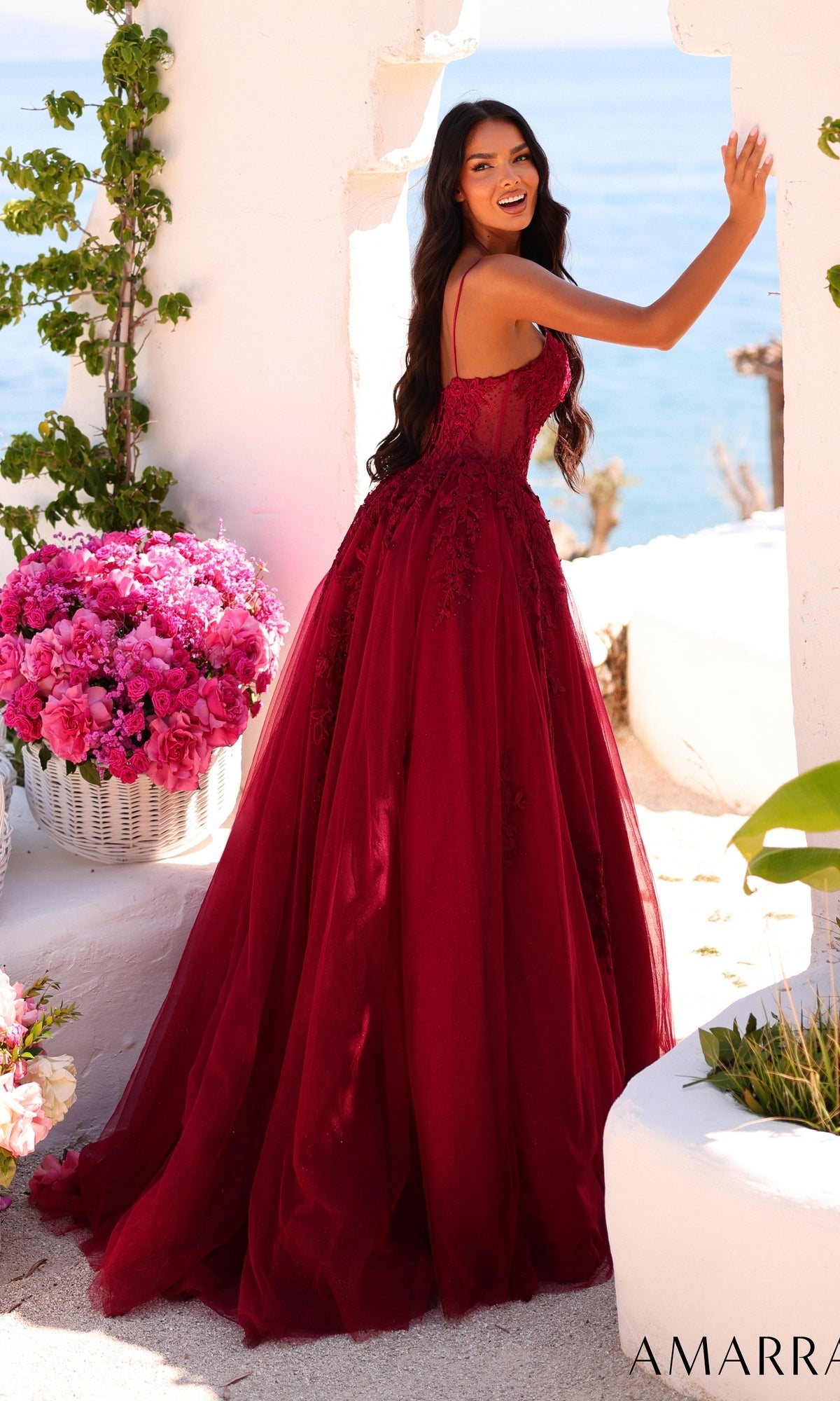 A woman wearing the Long Prom Dress: Amarra 89806—a sleeveless, deep red ball gown—stands outdoors by a wall, smiling. In the background are a large basket of pink flowers and the ocean.